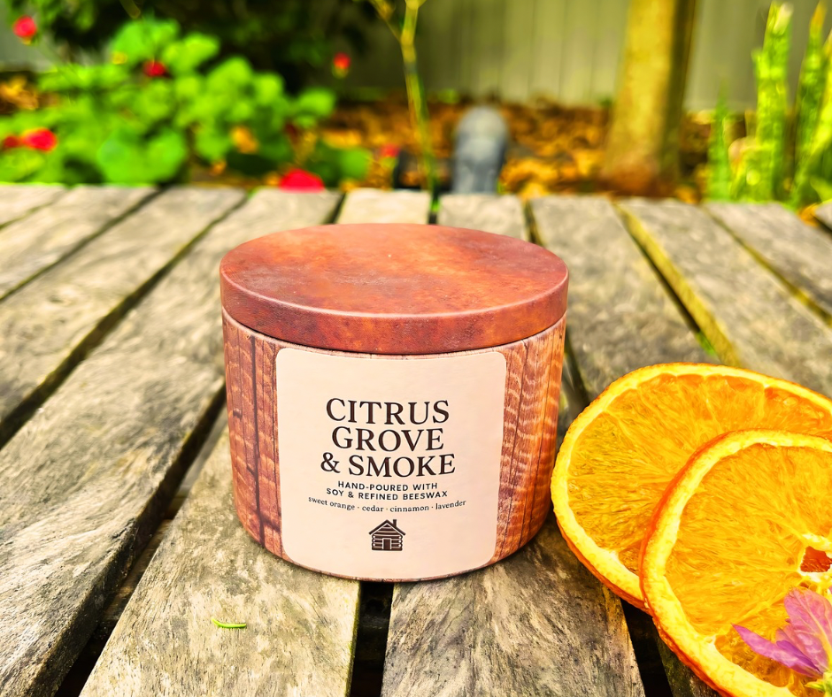 Candle labeled 'Citrus Grove & Smoke' on a wooden surface with orange slices and flowers in the background.