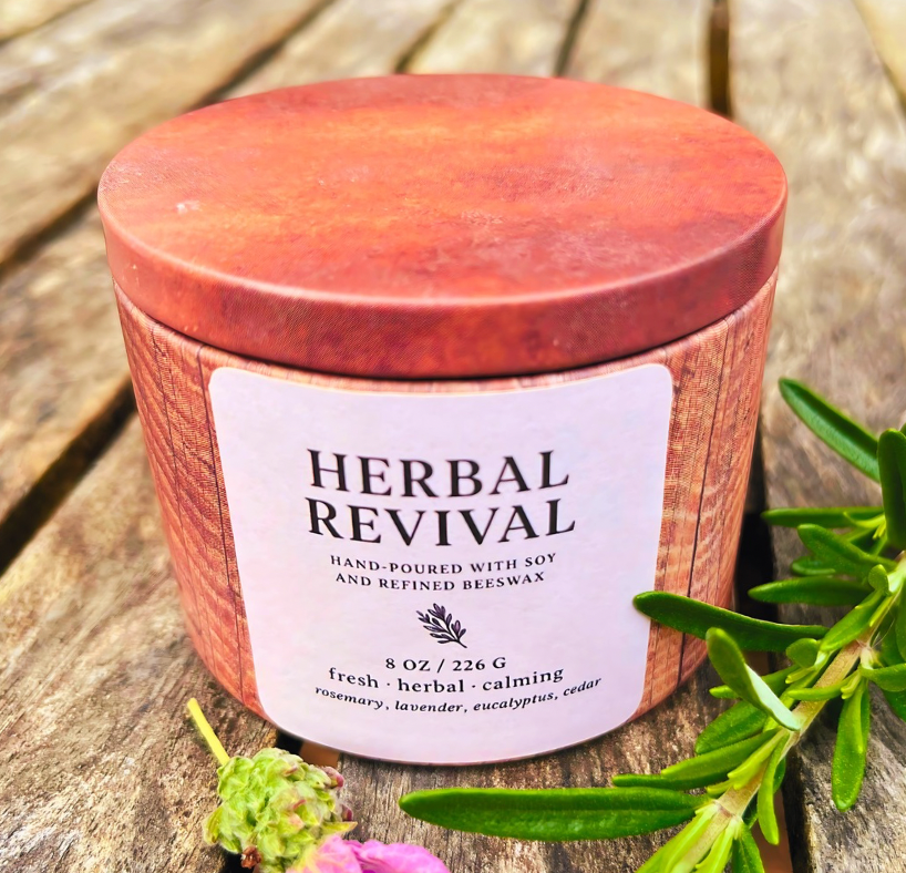 Candle labeled 'Herbal Revival' on a wooden surface with greenery.
