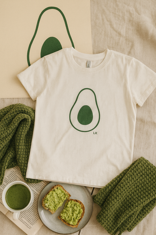 White t-shirt with avocado graphic on a beige surface with greenery
