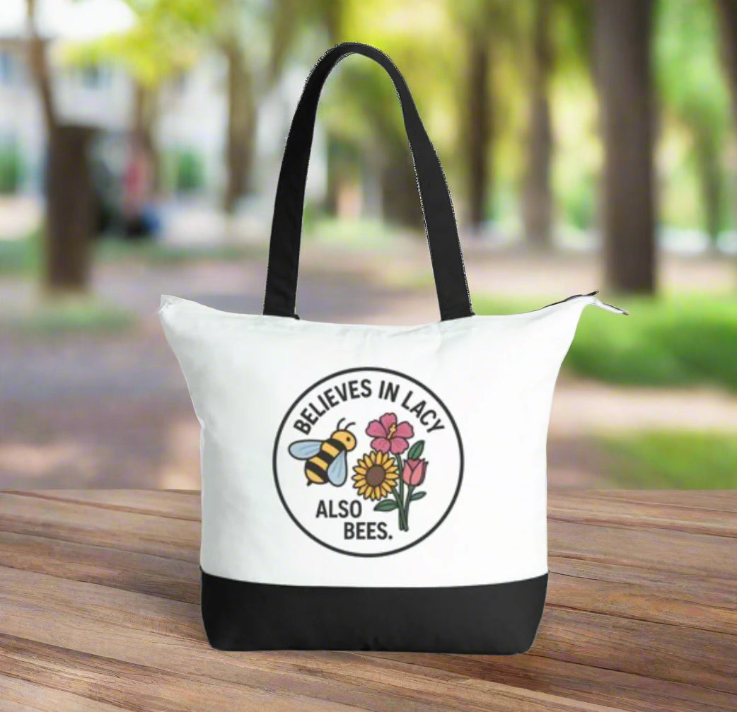 White tote bag with black handles and bottom section, featuring a bee and flower design with text.