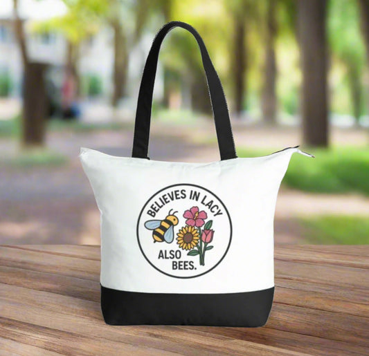 White tote bag with black handles and bottom section, featuring a bee and flower design with text.