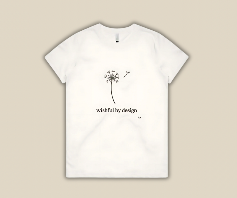 White t-shirt with a dandelion design and 'wishful by design' text on a beige background